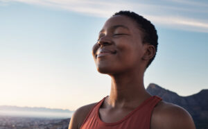 A Black woman in an orange tank top taking in a mindful breath, practicing mindfulness meditation with the sky as a background, symbolizing breaking free from ADHD.